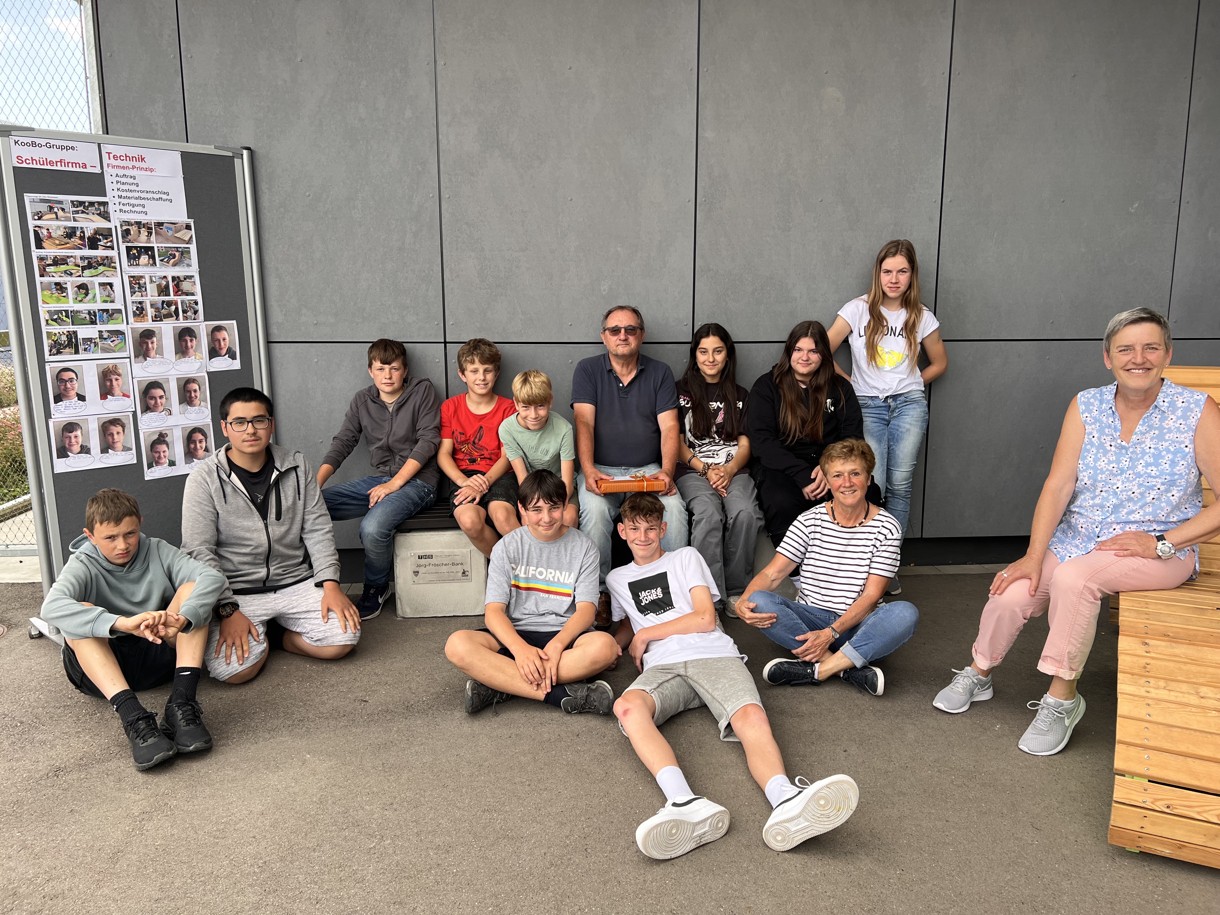 Bildbeschreibung: Schüler:innen sitzen mit Schulleiter Herrn Fröscher auf einer selbstgebauten Bank. Im Hintergrund steht eine Stellwand mit Fotos der Aktion.