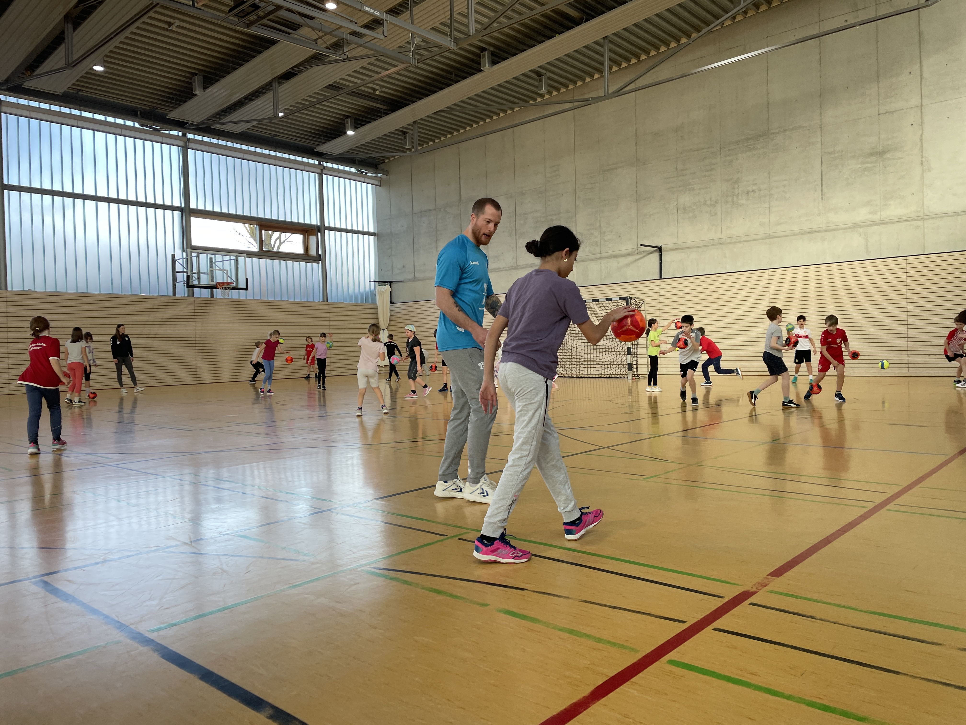 Im Vordergrund unterstützt ein Handballspieler ein Kind beim prellen. Im Hintergrund spielen Kinder mit dem Handball.