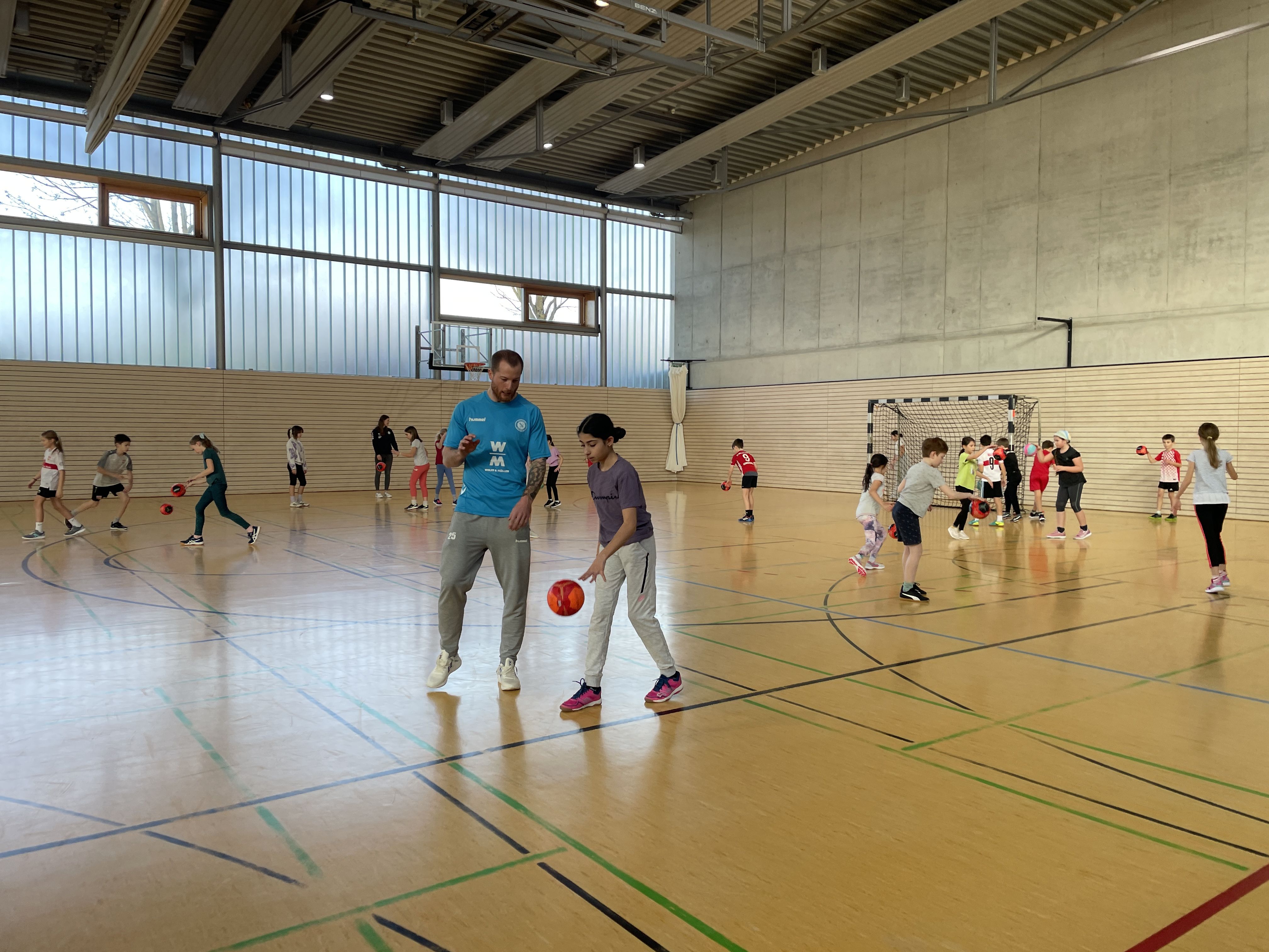 Im Vordergrund unterstützt ein Handballspieler ein Kind beim prellen. Im Hintergrund spielen Kinder mit dem Handball.