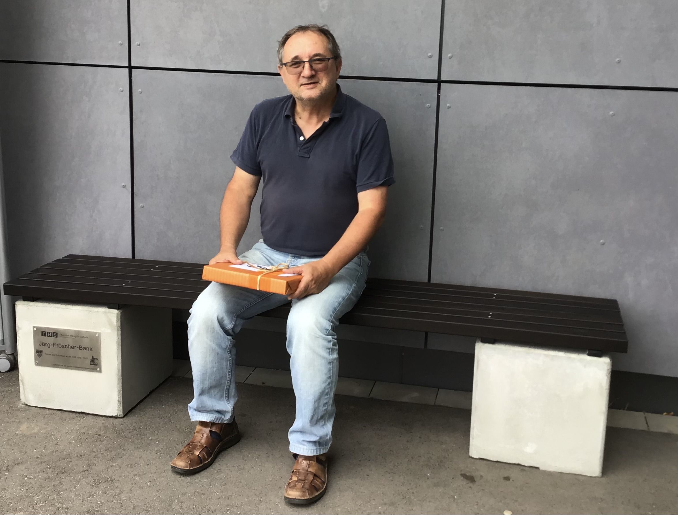Bildbeschreibung: Schulleiter Herr Fröscher sitzt auf einer selbstgebauten Bank mit "Jörg-Fröscher-Bank"-Plakette. Er hält ein Geschenk in der Hand.