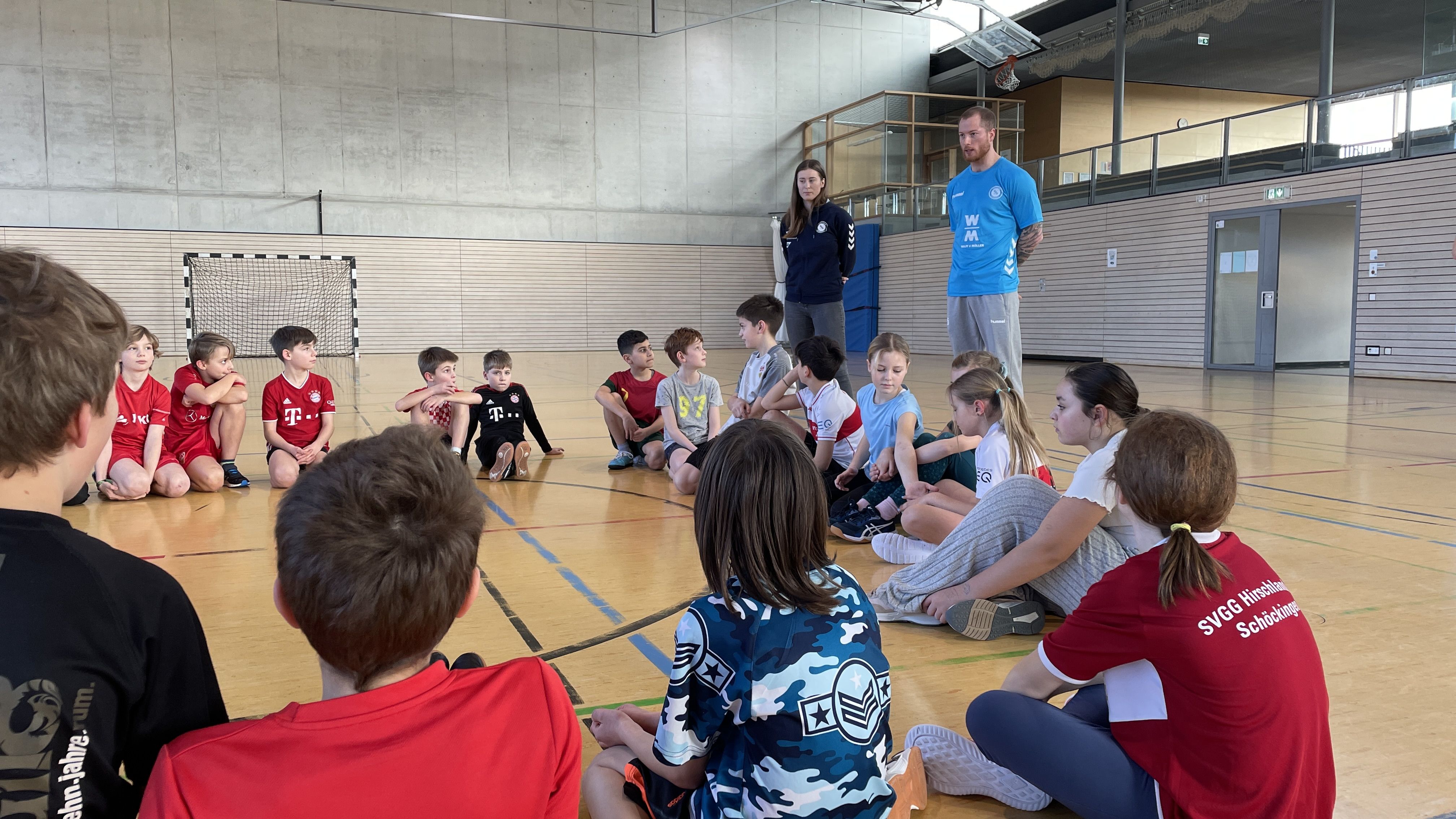 Kinder sitzen im Kreis in der Sporthalle und hören einem Handballspieler zu.