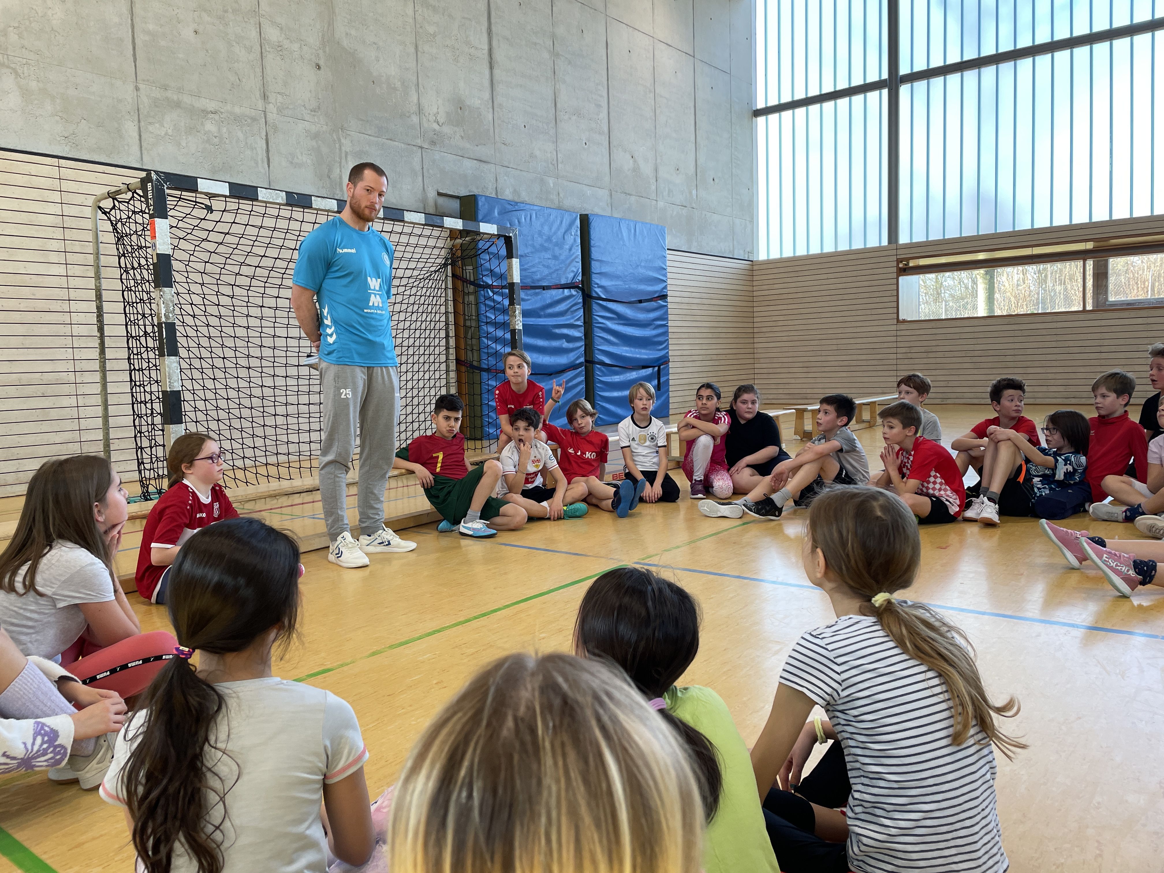 Kinder sitzen im Kreis am Handballtor, während der Handballspieler etwas erklärt.