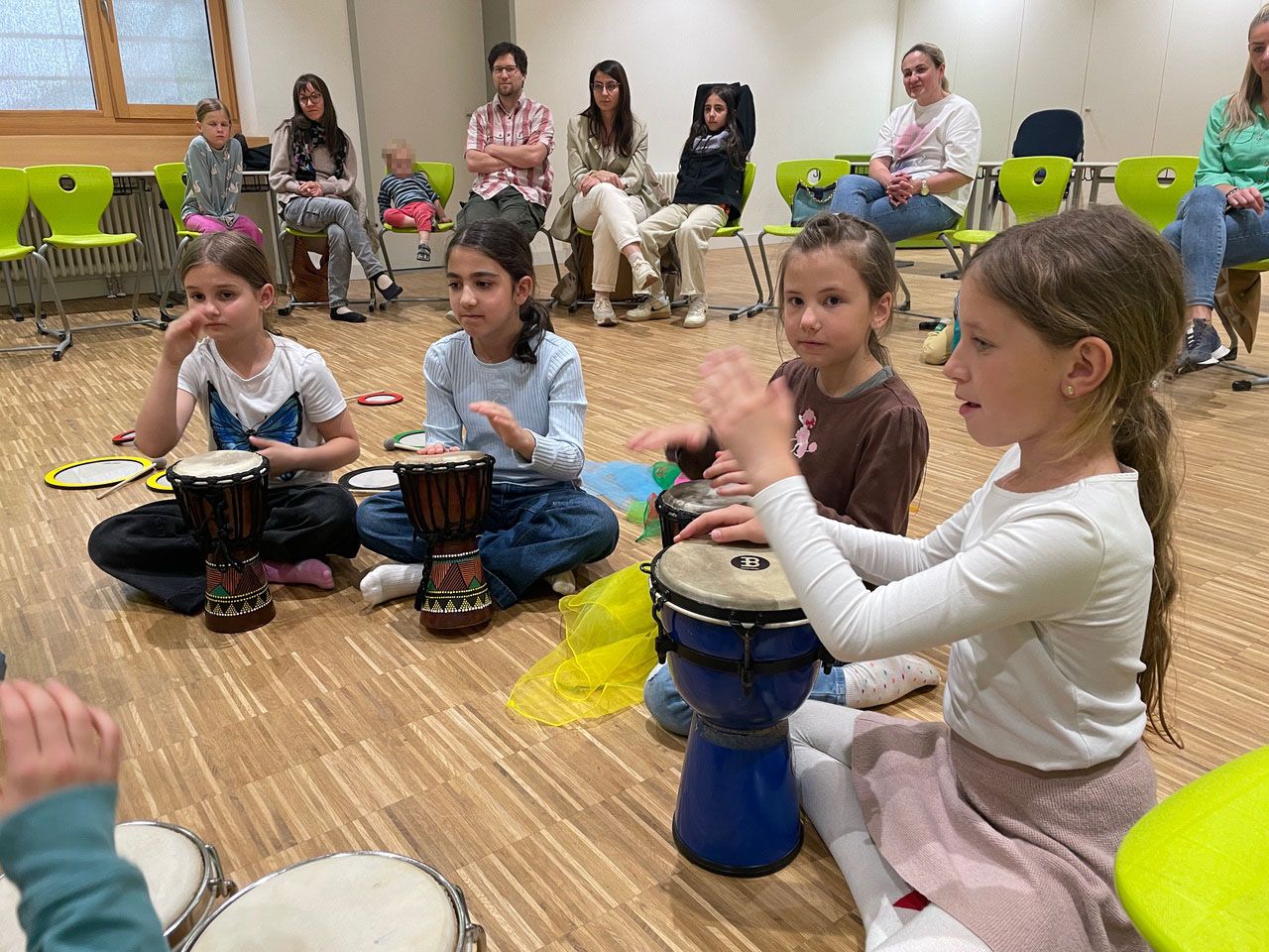 Mehrere Kinder spielen auf Trommeln. Dahinter sitzen Zuschauer auf Stühlen.