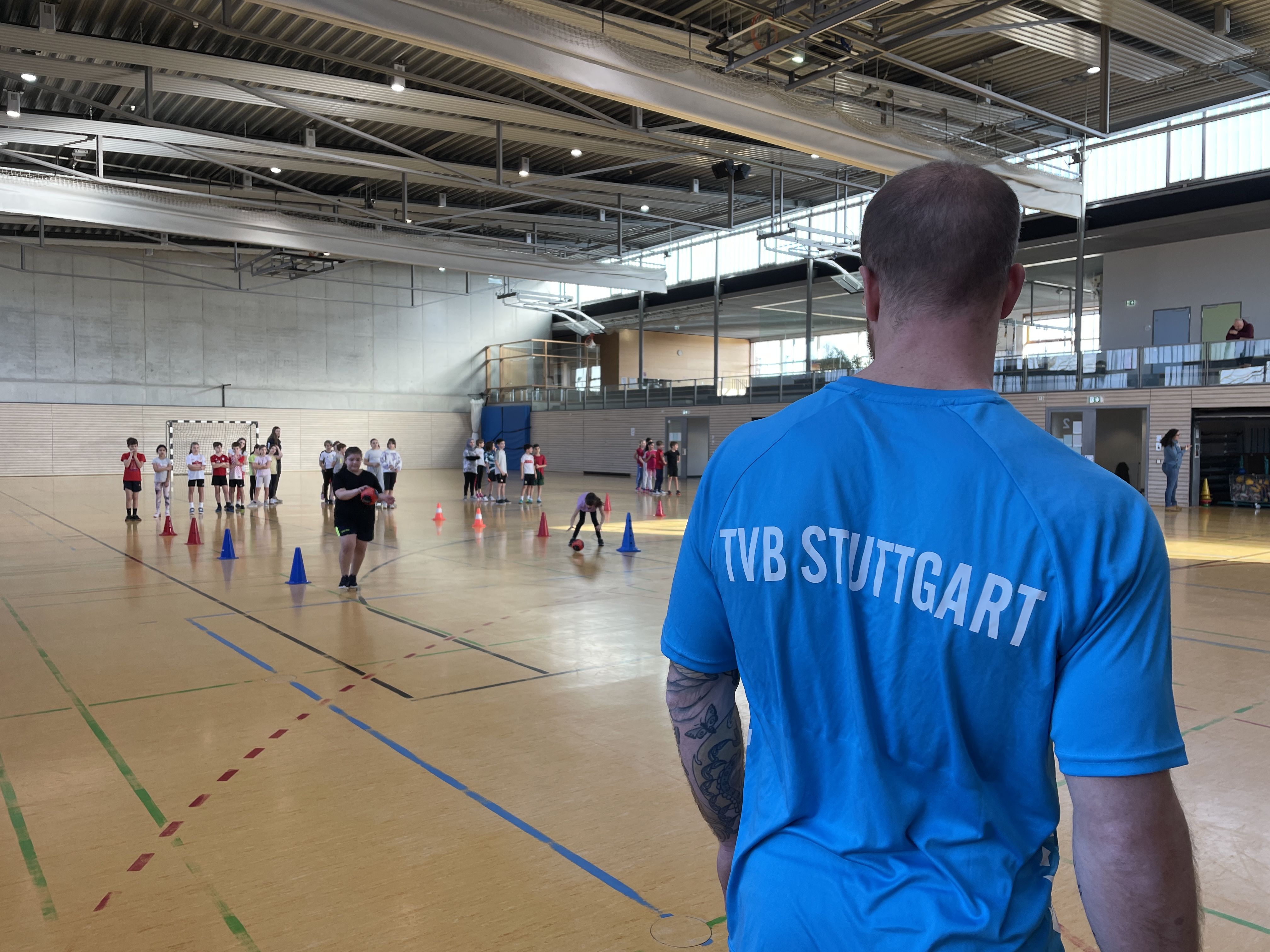 Im Vordergrund steht ein Handballspieler vom TVB Stuttgart und im Hintergrund machen Schüler eine Prellstaffel.